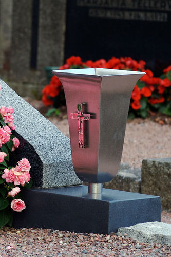 Hautakiven yhteydessä kiinteästi oleva metallinen, ristillä koristeltu kukkamaljakko.