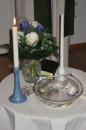 Kaksi kynttilää, kastemalja ja kukkaruukku pienellä pyöreällä pöydällä.