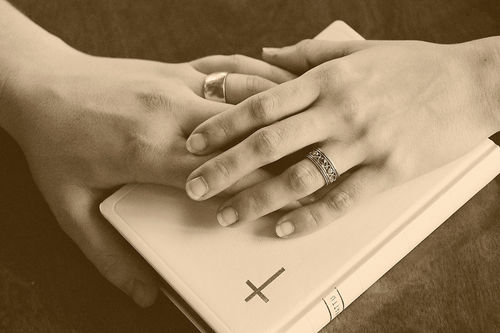 Kaksi päällekkäistä, sormustettua kättä Raamatun päällä.