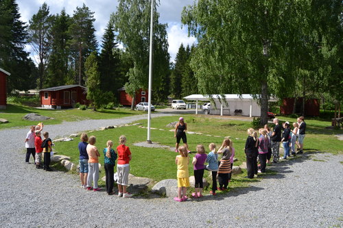 Joukko perheitä kokoontuneina leirinohjaajan ja lipputangon ympärille Kiikan Leiriaholla.